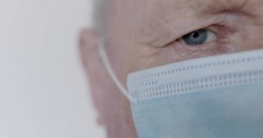 Arka planda tıbbi maskeli mavi gözlü kıdemli bir adamın portresi.