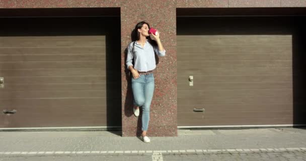 Стильная девушка стоит в позе на парковке, пьет кофе и радуется — стоковое видео