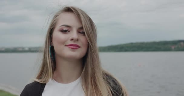 Chica alegre corrige su pelo que sopla y sonríe a la cámara en el paseo marítimo — Vídeos de Stock