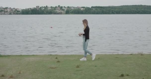 Gadis ceria berjalan di tepi sungai, mengoreksi rambutnya yang bertiup dan tersenyum — Stok Video