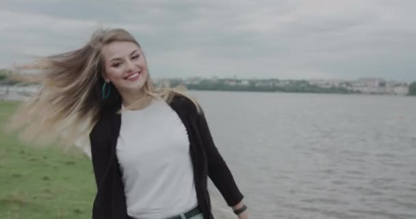 Gadis bahagia dengan rambut bertiup dan pakaian santai berjalan di tepi sungai dan berpose — Stok Video