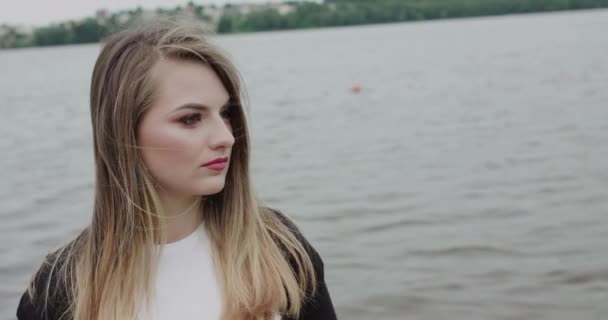 Задумчивый взгляд на камеру красивой девушки с развевающимися волосами, стоящей у реки — стоковое видео