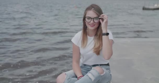 Счастливая девушка расслабляется на реке в ветреный день, корректирует очки и оглядывается вокруг — стоковое видео