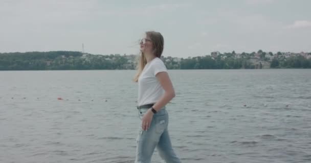 楽しいです女の子で眼鏡で歩く川沿いとポーズでカメラで風の強い日 — ストック動画