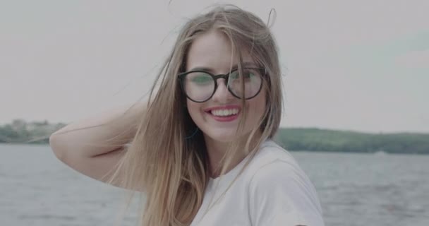 Fille pose à la caméra, corrige les cheveux longs, soufflant et souriant à la rivière — Video