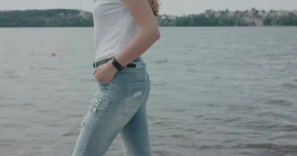 Dívka kráčí na břehu řeky a otáčí se s foukající vlasy na kameru — Stock video