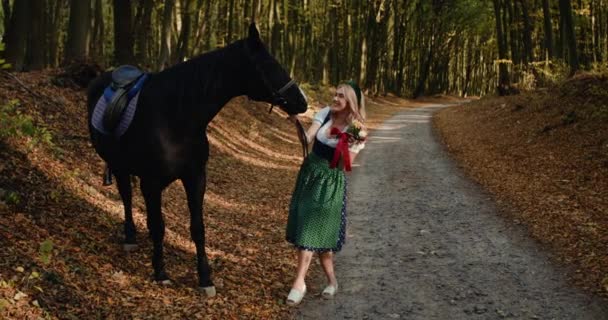 Красивая блондинка с букетом диких цветов, позирует с лошадью в лесу — стоковое видео
