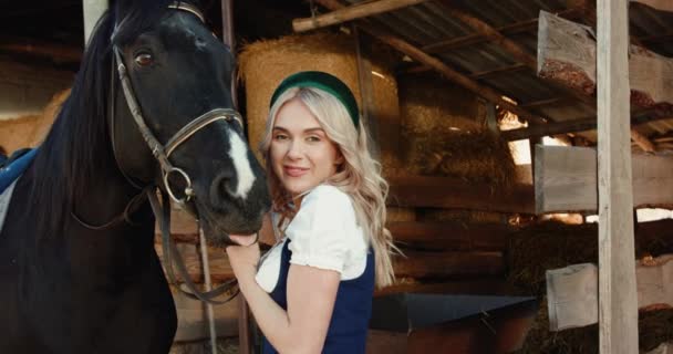 Rubia sonriendo y acariciando un lindo negro caballos cabeza a cámara en estable — Vídeo de stock