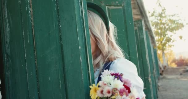 Meisje met boeket bloemen kijkt uit de staldeur en flirt met de camera — Stockvideo