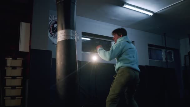 Sam bokser uderza w worek treningowy w ciemnej sali gimnastycznej w zwolnionym tempie. Młody człowiek trenuje w domu. Silny atleta na siłowni. Koncepcja sportu. — Wideo stockowe