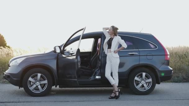 Beautiful lady in white suit and black shoes poses at car out of city — Stock Video