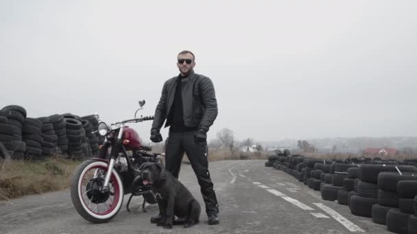 Potrait de motorista brutal se para en moto, neumáticos y bulldog oscuro — Vídeo de stock