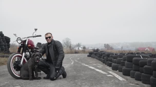 Potrait de motociclista feliz sentado en la motocicleta, neumáticos y sostiene bulldog oscuro — Vídeos de Stock