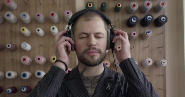 Hombre guapo escuchando música y sonriendo a la cámara en el atelier en el conjunto de hilos — Vídeo de stock