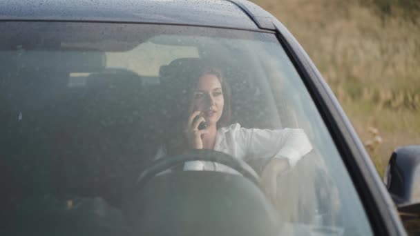 Перегляд через мокрий з дощем вітрове скло усміхненої дівчини в машині розмовляє по телефону — стокове відео
