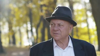 Kıdemli bir adamın şapkalı portresi kenara bakar ve sonbaharda, güneşli parkta hayranlık duyar.