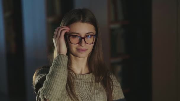 Menina com óculos tucks seu cabelo atrás de sua orelha e ri da câmera na biblioteca — Vídeo de Stock