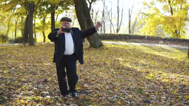 Homem sênior se divertindo e dançando em um parque de outono ao ouvir música — Vídeo de Stock