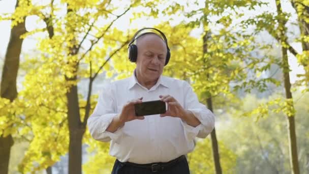 Senior en camisa se mueve cabeza asides cuando se ve vídeo en el teléfono con auriculares — Vídeos de Stock