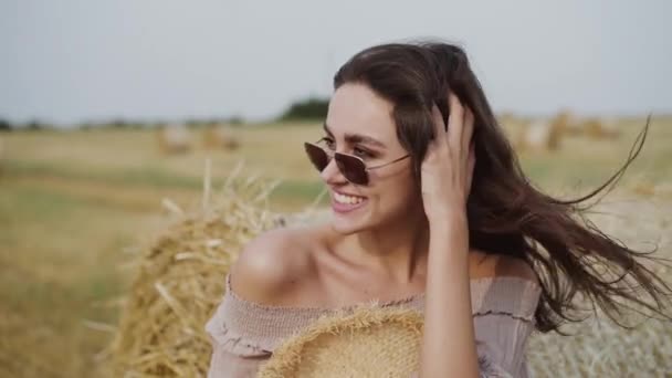 Femme heureuse se réjouit à la meule de foin et corrige les cheveux pendant le vent fort dans le champ — Video
