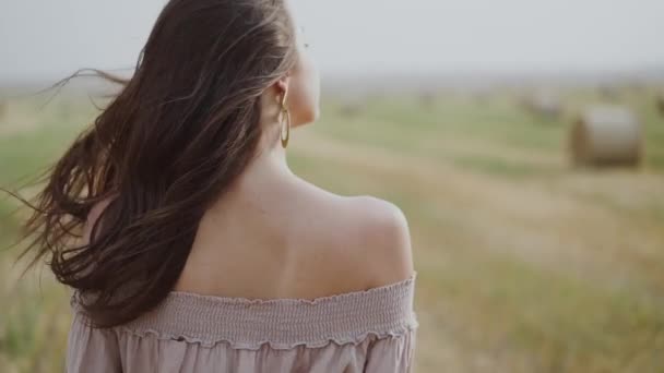 Ragazza cammina tra il campo di paglia ventoso, si gira verso la fotocamera, si mette il cappello e sorride — Video Stock