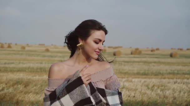 Красива дівчина з пледом радіє і позує у вітряному полі зі стопами сіна — стокове відео