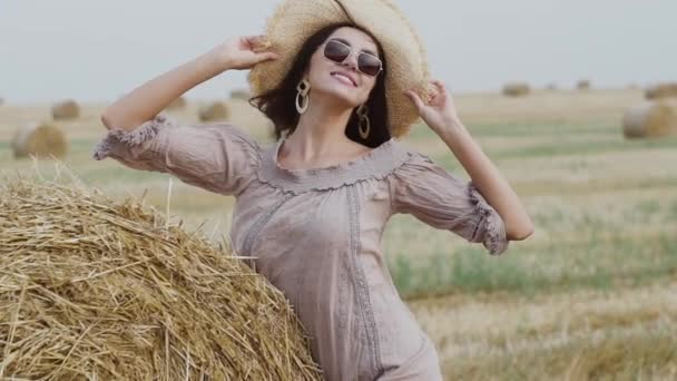 Элегантная брюнетка в солнцезащитных очках стоит в позе у стога сена и надевает шляпу — стоковое видео