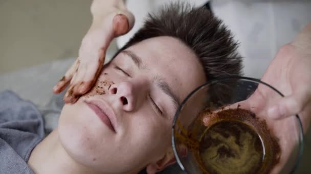 Beauticians hand applies coffee scrub on mans face skin with massage movements — Stock Video