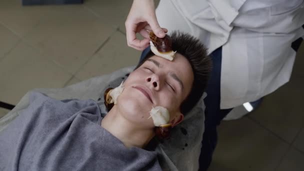 Ręce kosmetyczek stosujących pełzające ślimaki na twarzy człowieka do odmłodzenia skóry — Wideo stockowe