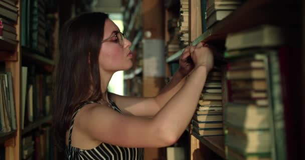 Estudiante mira libros en la biblioteca, saca uno y lo mira con alegría — Vídeos de Stock