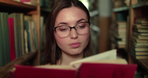 Porträt eines Mädchens mit Brille, das das Buch vor der Kamera zwischen Regalen in der Bibliothek liest — Stockvideo