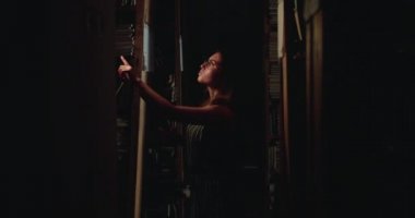 Gözlüklü bir kız karanlıkta kütüphane raflarında kitap arıyor.