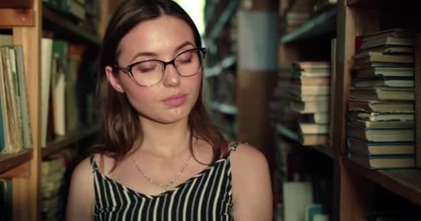 Девушка листает книгу и с радостью смотрит на нее среди полок в библиотеке — стоковое видео