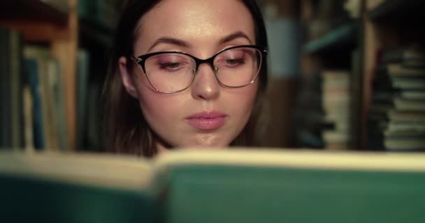 Близкий портрет девушки в очках читает книгу у камеры на полках в библиотеке — стоковое видео