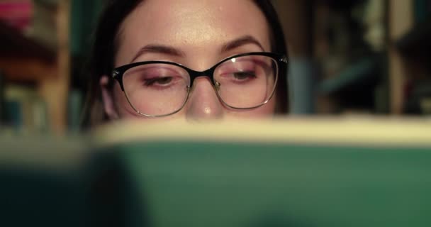 Близкий портрет девушки в очках, читающей книгу, ведет глаза к камере в библиотеке — стоковое видео