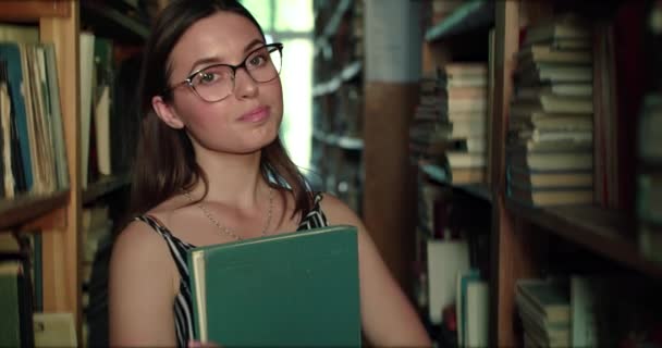Девушка в очках с книгой в руках, слегка улыбающаяся перед камерой в библиотеке — стоковое видео