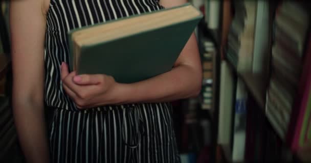 Chica irreconocible sostiene el libro en las manos entre los estantes de la biblioteca — Vídeos de Stock