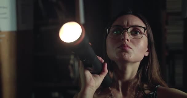 Chica seria en gafas con una linterna ve los estantes de la biblioteca en la oscuridad — Vídeos de Stock