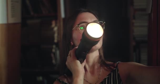 Chica con linterna ve estantes de la biblioteca en la oscuridad, se sorprende y tira de libro — Vídeos de Stock