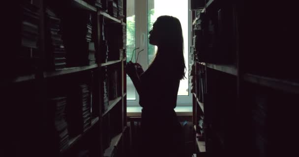 Жіночий силует, одягаючи окуляри та переглядаючи книги на полицях бібліотеки — стокове відео