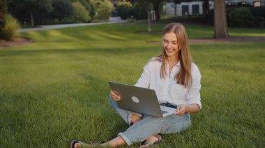 Genç bir öğrenci çimlerde dizüstü bilgisayarla otururken çalışıyor. 4k