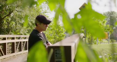 Köprü yapraklı parka dayanan adam, yeşil yaprakların arasından süzülen yumuşak güneş ışığı, kepli rahat duruş, ahşap korkuluklara yaslanma, sığ odak, yaz mevsimi