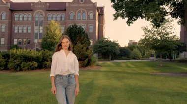 Genç beyaz kadın gün batımında kampüs çimlerinde duruyor. Arka planda tarihi tuğla üniversite binası, sıradan kot pantolonlar.