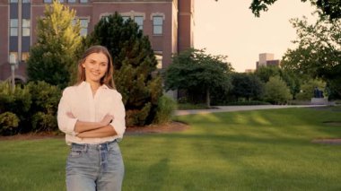 Kampüs kollarında çapraz duran genç beyaz kadın, kot pantolon ve beyaz bluz giyiyor, yeşil çimlerin ve ağaçların üzerinde yumuşak altın güneş ışığı,