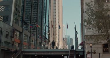 Chicago şehir merkezinde üst geçitte yayalar var. Yükselen gökdelenler, dalgalanan bayraklar ve köprüden geçen siluetli banliyöler tarafından çerçevelenmiş.