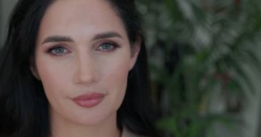 Makyajlı güzel bir esmer kadın kameraya bakıyor, gülümsüyor, arka planda yeşil bitkiler var. Yavaş çekim. 4K