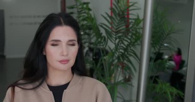 Makyajlı güzel bir esmer kadın gözlerini açıyor ve kameraya bakıyor, gülümsüyor, arka planda yeşil bitkiler var. 4K