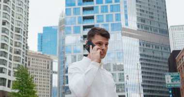 İş dünyası tarzında genç bir adamın, büyük bir iş bölgesindeki yüksek binaların arka planında telefonla konuştuğu yavaş çekim. 4K