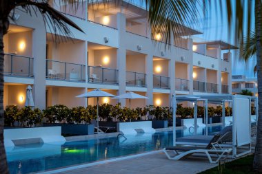 Aydınlatılmış balkonları ve alacakaranlıkta yüzme havuzu olan modern bir otel binası.