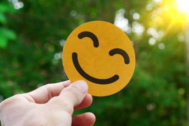 Güneşli, yeşil bir arka plana karşı sarı göz kırpan bir gülen yüz ikonu tutan bir insan eli.